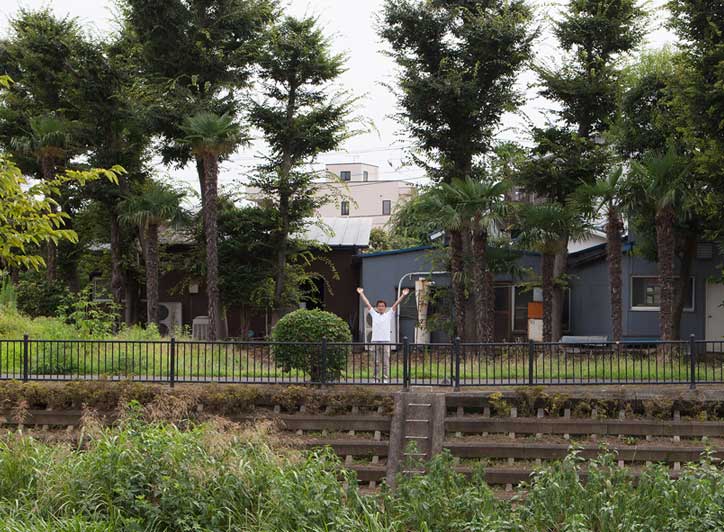 背の高いヤシの木が並ぶ庭で、両手を広げて立つ白い服の関太郎（せき たろう）。野川のほとりで鉄柵と石垣に囲まれた緑豊かな木工所。