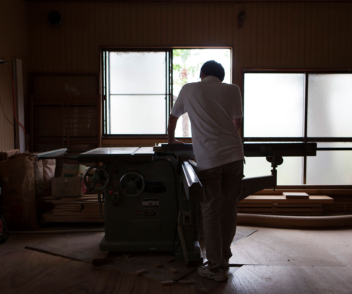 木工室で大型の工作機械に向かう作業員の関太郎（せき たろう）のシルエット。窓からの自然光が室内を照らしている。