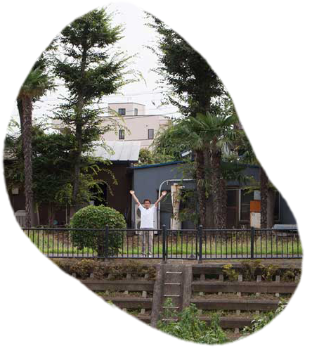 背の高いヤシの木が並ぶ庭で、両手を広げて立つ白い服の関太郎（せき たろう）。野川のほとりで鉄柵と石垣に囲まれた緑豊かな木工所。